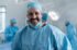 Smiling caucasian male surgeon with face mask and protective clothing in operating theatre. medicine, health and healthcare services during covid 19 coronavirus pandemic.