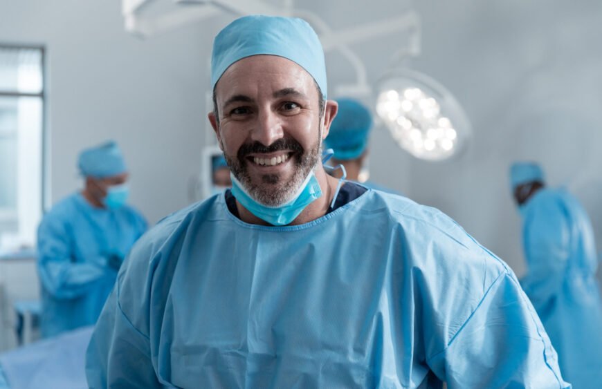 Smiling caucasian male surgeon with face mask and protective clothing in operating theatre. medicine, health and healthcare services during covid 19 coronavirus pandemic.